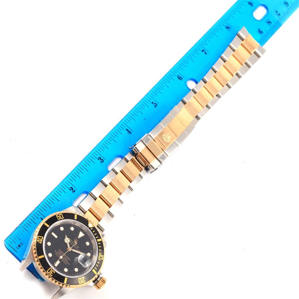 The image shows a Rolex Submariner 16613 Men's Steel and Gold (two tone) Black Dial 16613 Men's Steel and Gold (two tone) Black Dial watch placed horizontally beside a blue ruler, highlighting its black dial, bezel, and gold and silver bracelet.