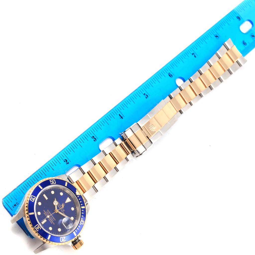 The image shows a Rolex Submariner 16613 Men's Steel and Gold (two tone) Blue Dial 16613 Men's Steel and Gold (two tone) Blue Dial watch laid flat with the bracelet and bezel visible, next to a blue ruler.
