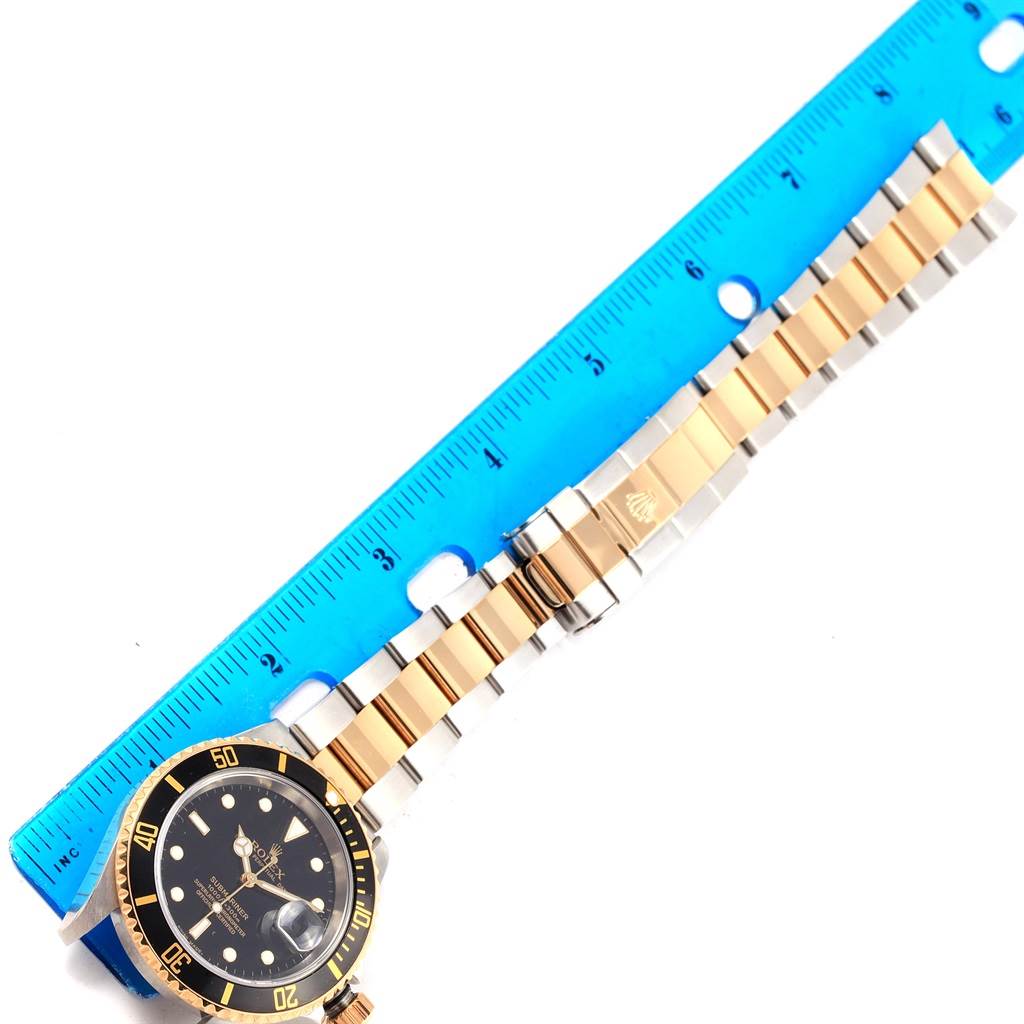 The image shows a Rolex Submariner 16613 Men's Steel and Gold (two tone) Black Dial 16613 Men's Steel and Gold (two tone) Black Dial watch laid flat against a ruler, revealing the face, bezel, and bracelet.