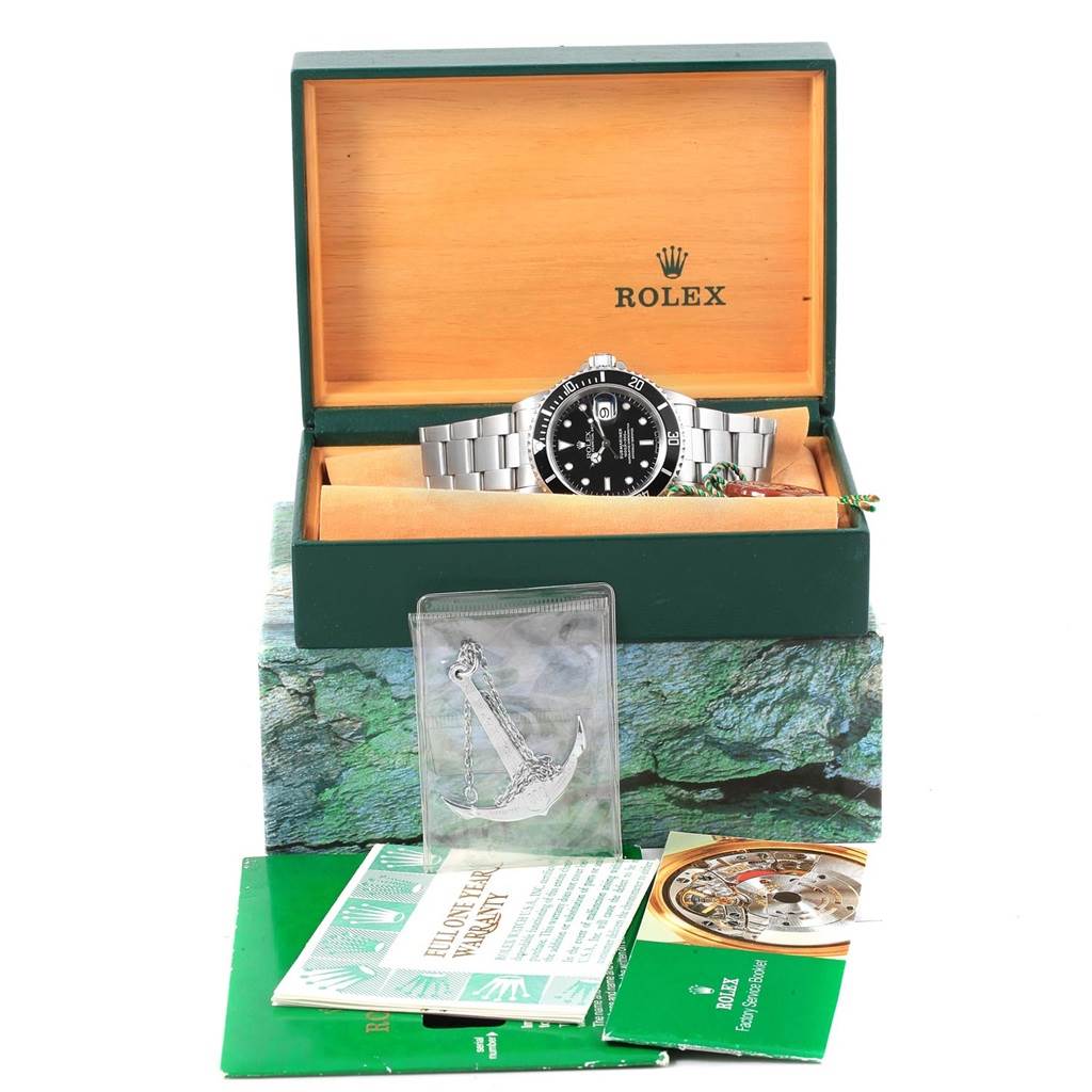 The Rolex Submariner 16610 Men's Stainless Steel Black Dial 16610 Men's Stainless Steel Black Dial watch is shown at a front angle in an open box, with accompanying warranty booklets and an accessory.