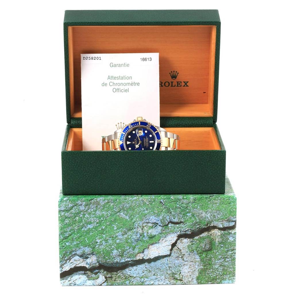 The Rolex Submariner 16613 Men's Steel and Gold (two tone) Blue Dial 16613 Men's Steel and Gold (two tone) Blue Dial watch is shown in its open green box with documentation in the background.