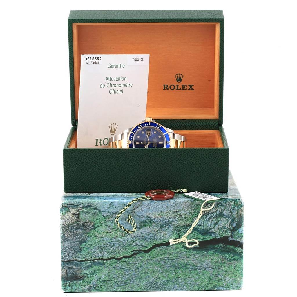 The Rolex Submariner 16613 Men's Steel and Gold (two tone) Blue Dial 16613 Men's Steel and Gold (two tone) Blue Dial watch is displayed front-facing in an open box, along with its warranty and chronometer certification.