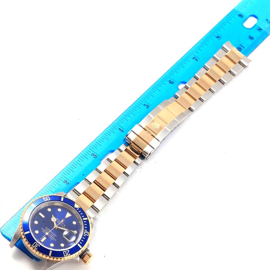 The image shows a Rolex Submariner 16613 Men's Steel and Gold (two tone) Blue Dial 16613 Men's Steel and Gold (two tone) Blue Dial watch with a blue bezel and dial, positioned beside a blue ruler for measurement.