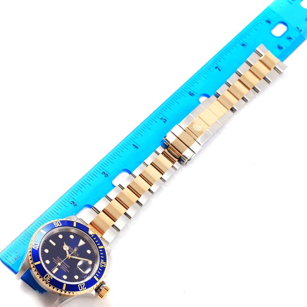 The Rolex Submariner 16613 Men's Steel and Gold (two tone) Blue Dial 16613 Men's Steel and Gold (two tone) Blue Dial watch is shown from a top-down angle, displaying the face, bezel, and bracelet, beside a blue ruler.