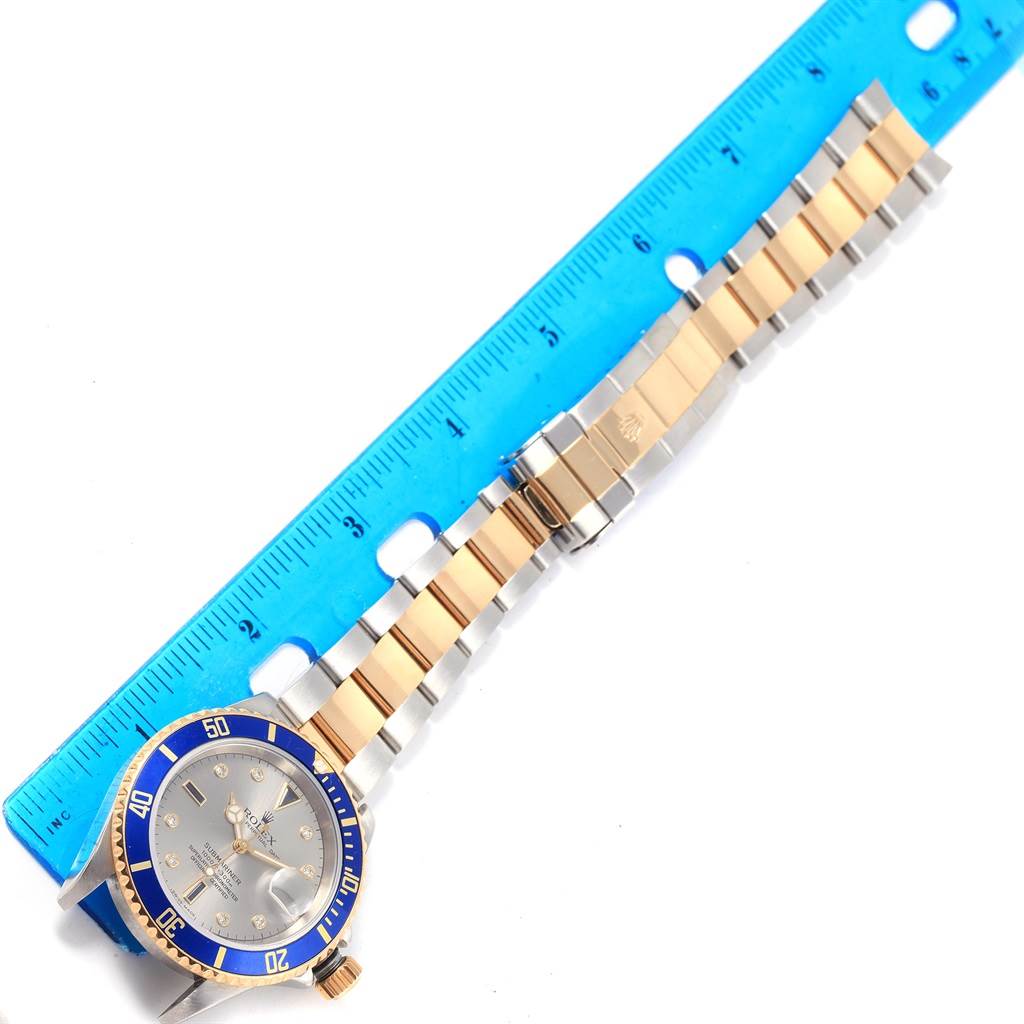 The image shows a Rolex Submariner 16613 Men's Steel and Gold (two tone) Grey Dial 16613 Men's Steel and Gold (two tone) Grey Dial watch laid flat next to a blue ruler, displaying its face and two-tone gold and silver bracelet.