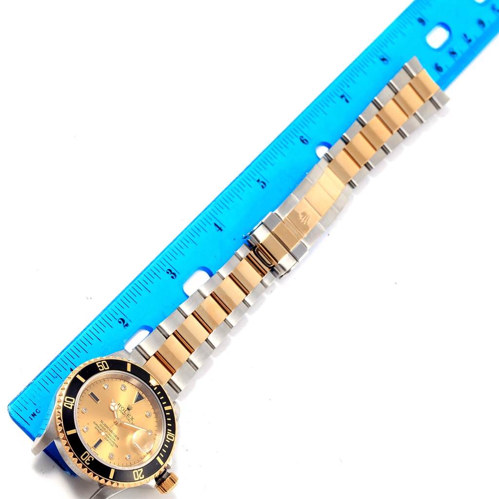 The image shows a Rolex Submariner 16613 Men's Steel and Gold (two tone) Champagne Dial 16613 Men's Steel and Gold (two tone) Champagne Dial watch laid flat with its gold and silver bracelet extended alongside a blue ruler.