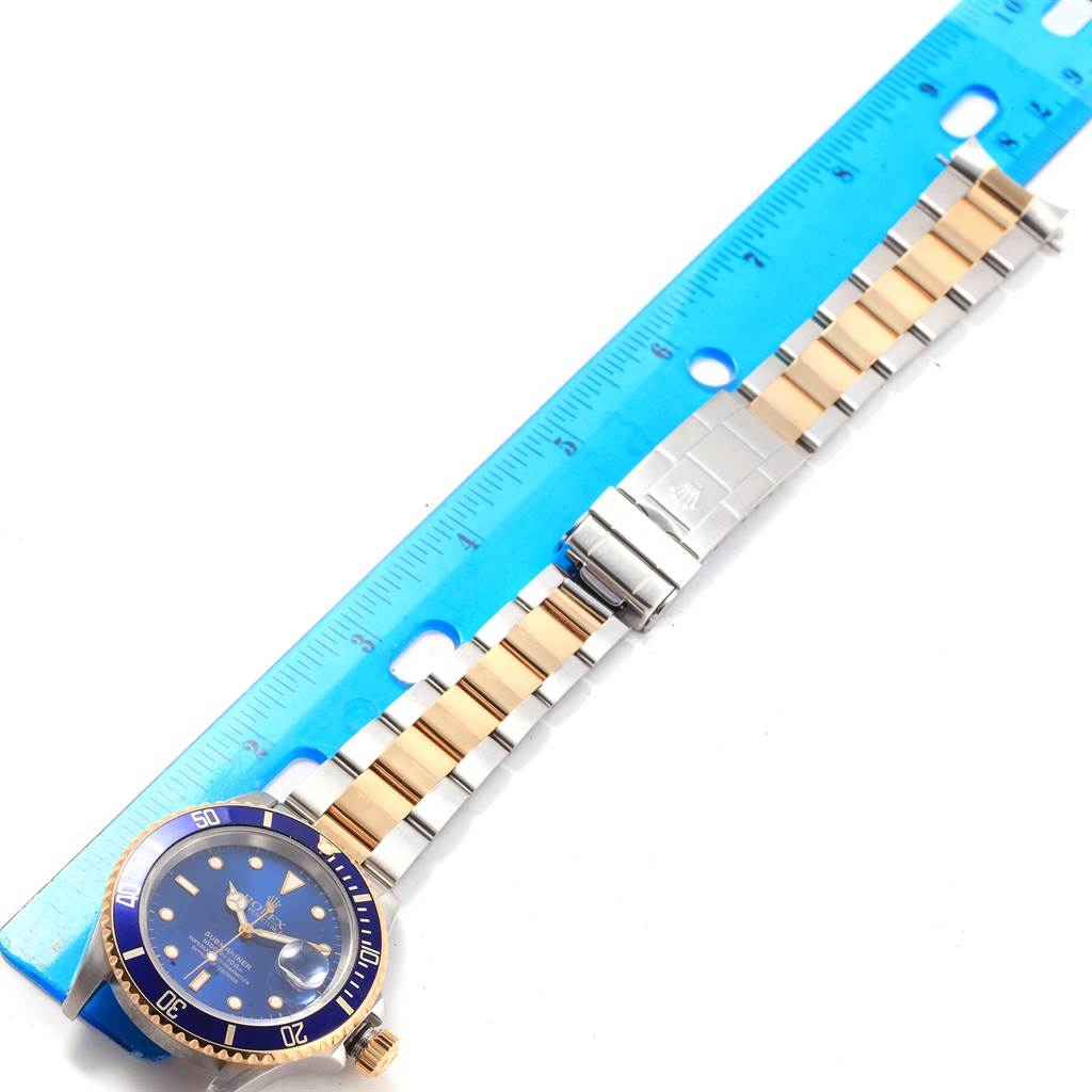 The image shows a Rolex Submariner 16613 Men's Steel and Gold (two tone) Blue Dial 16613 Men's Steel and Gold (two tone) Blue Dial watch laid flat, including the face, bezel, bracelet, and clasp next to a blue ruler.