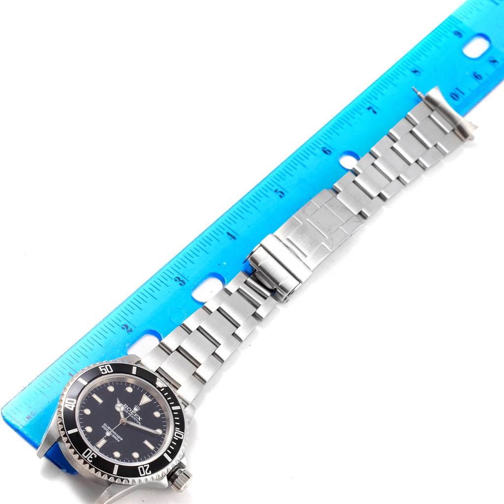 The image shows a Rolex Submariner 14060 Men's Stainless Steel Black Dial 14060 Men's Stainless Steel Black Dial watch with an outstretched stainless steel bracelet next to a blue ruler.