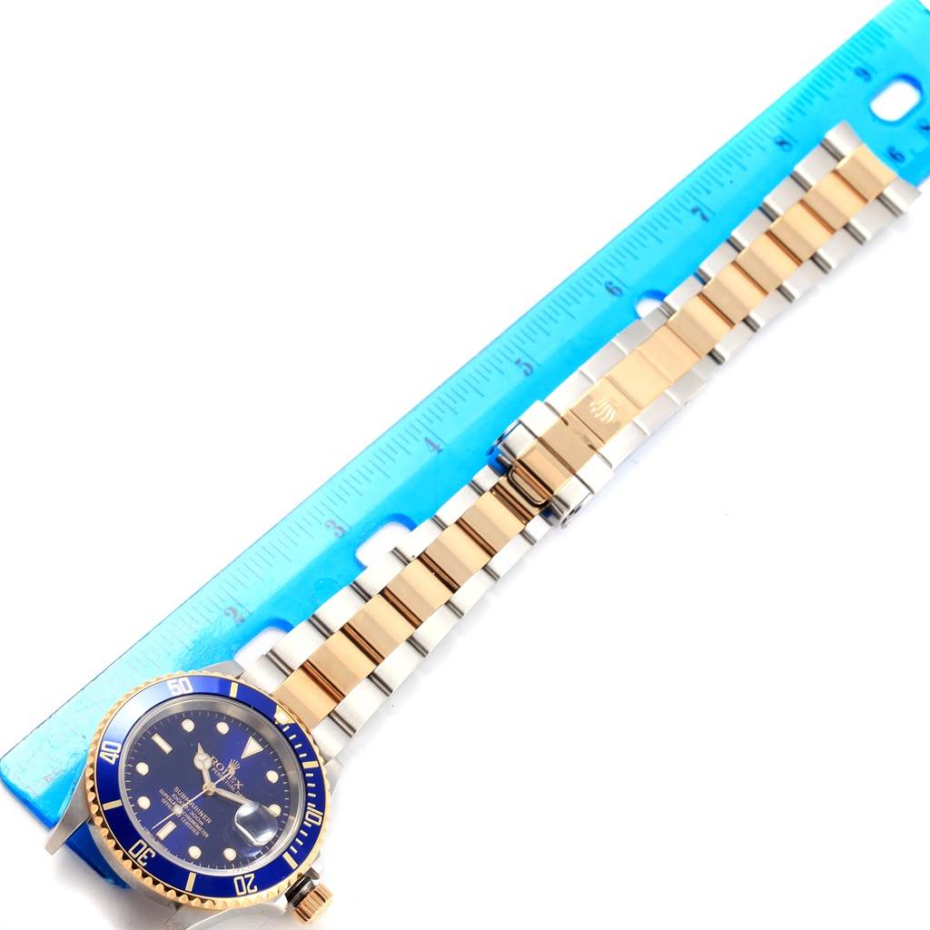 The image shows a Rolex Submariner 16613 Men's Steel and Gold (two tone) Blue Dial 16613 Men's Steel and Gold (two tone) Blue Dial watch laid flat along a blue ruler, displaying its case, bezel, dial, and bracelet.
