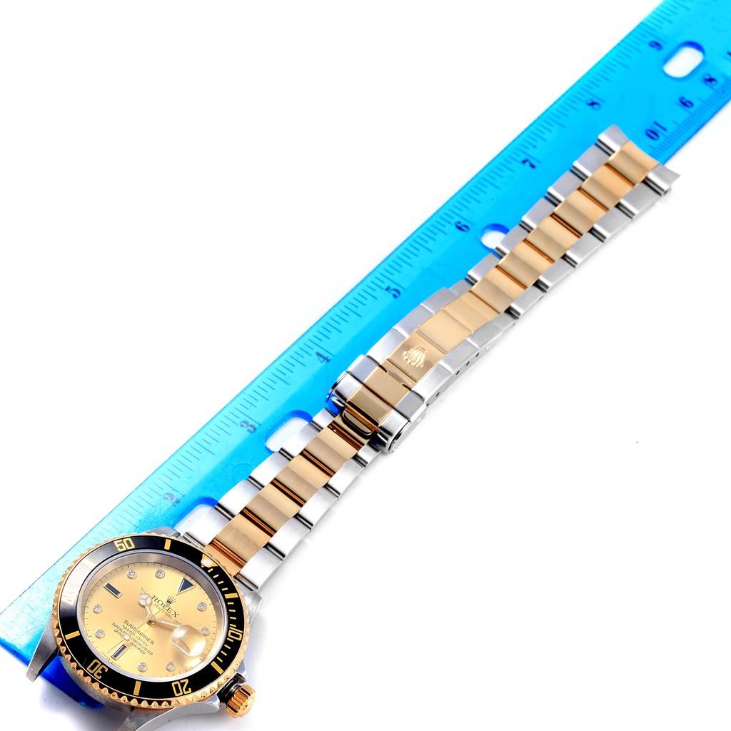 The image shows a Rolex Submariner 16613 Men's Steel and Gold (two tone) Champagne Dial 16613 Men's Steel and Gold (two tone) Champagne Dial watch face-up next to a ruler, revealing the bezel, face, crown, and bracelet.