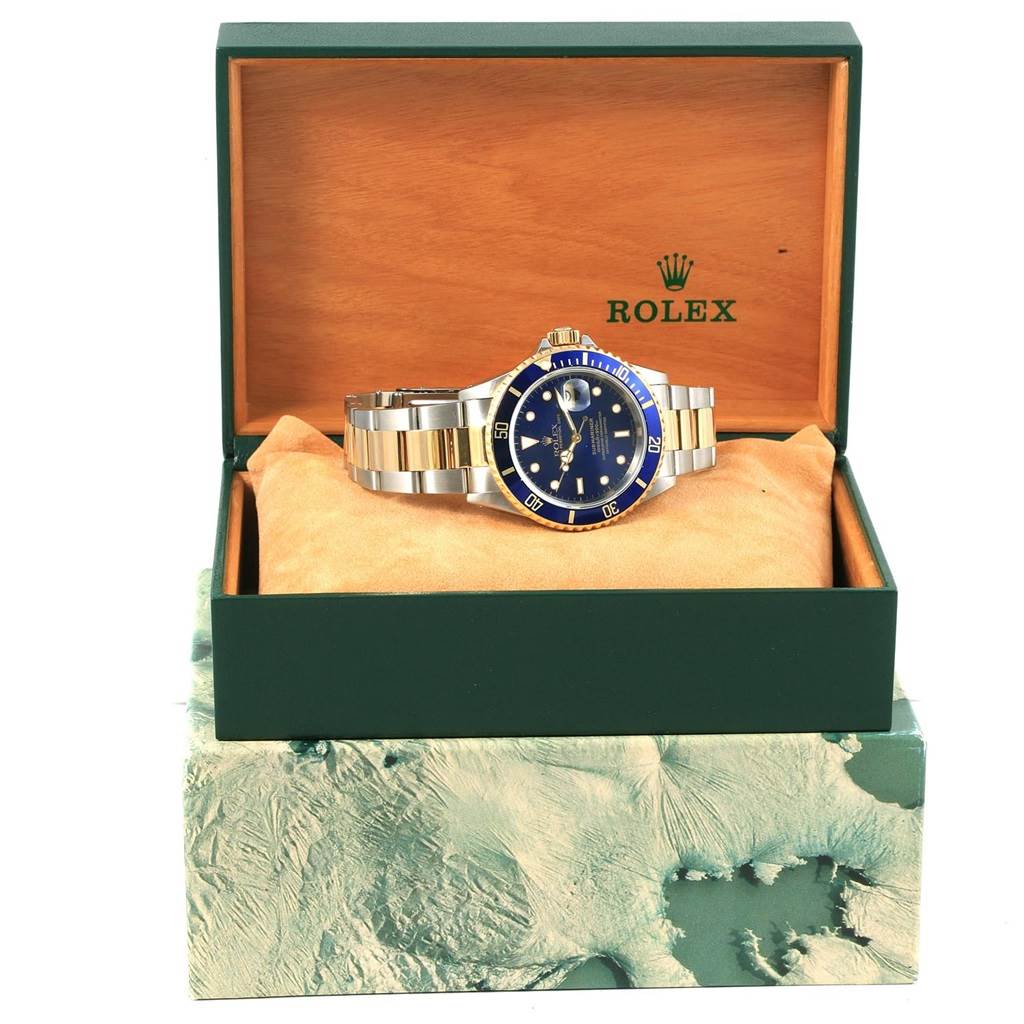 The image shows a Rolex Submariner 16613 Men's Steel and Gold (two tone) Blue Dial 16613 Men's Steel and Gold (two tone) Blue Dial watch with a blue dial and gold accents, displayed at an angle inside an open green Rolex box.