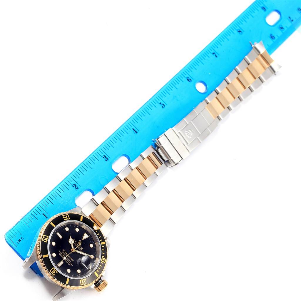 The image shows a Rolex Submariner 16613 Men's Steel and Gold (two tone) Black Dial 16613 Men's Steel and Gold (two tone) Black Dial watch laid flat with the face and bracelet visible alongside a blue ruler.