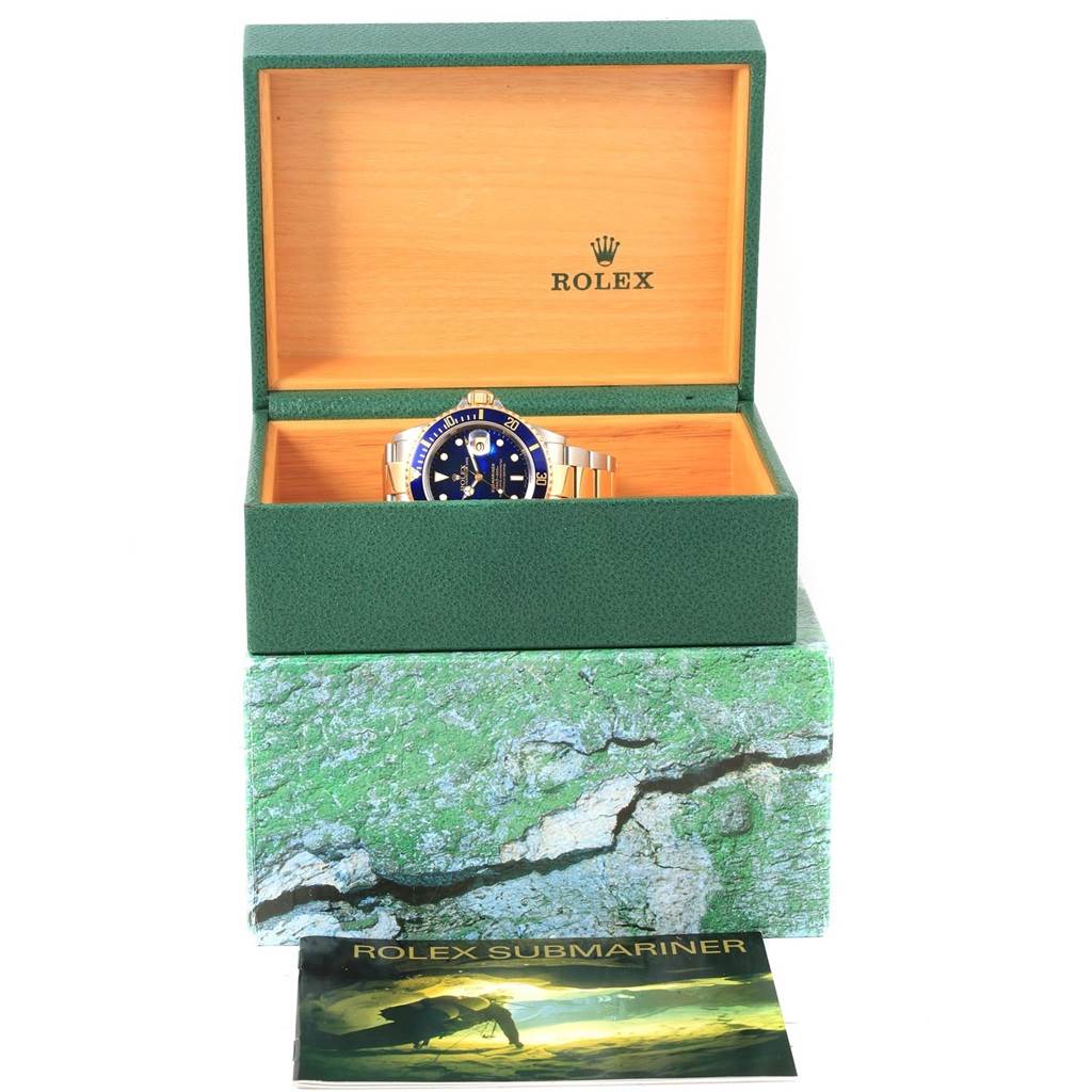 The Rolex Submariner 16613 Men's Steel and Gold (two tone) Blue Dial 16613 Men's Steel and Gold (two tone) Blue Dial watch is displayed in its open green box, with a booklet placed below it.