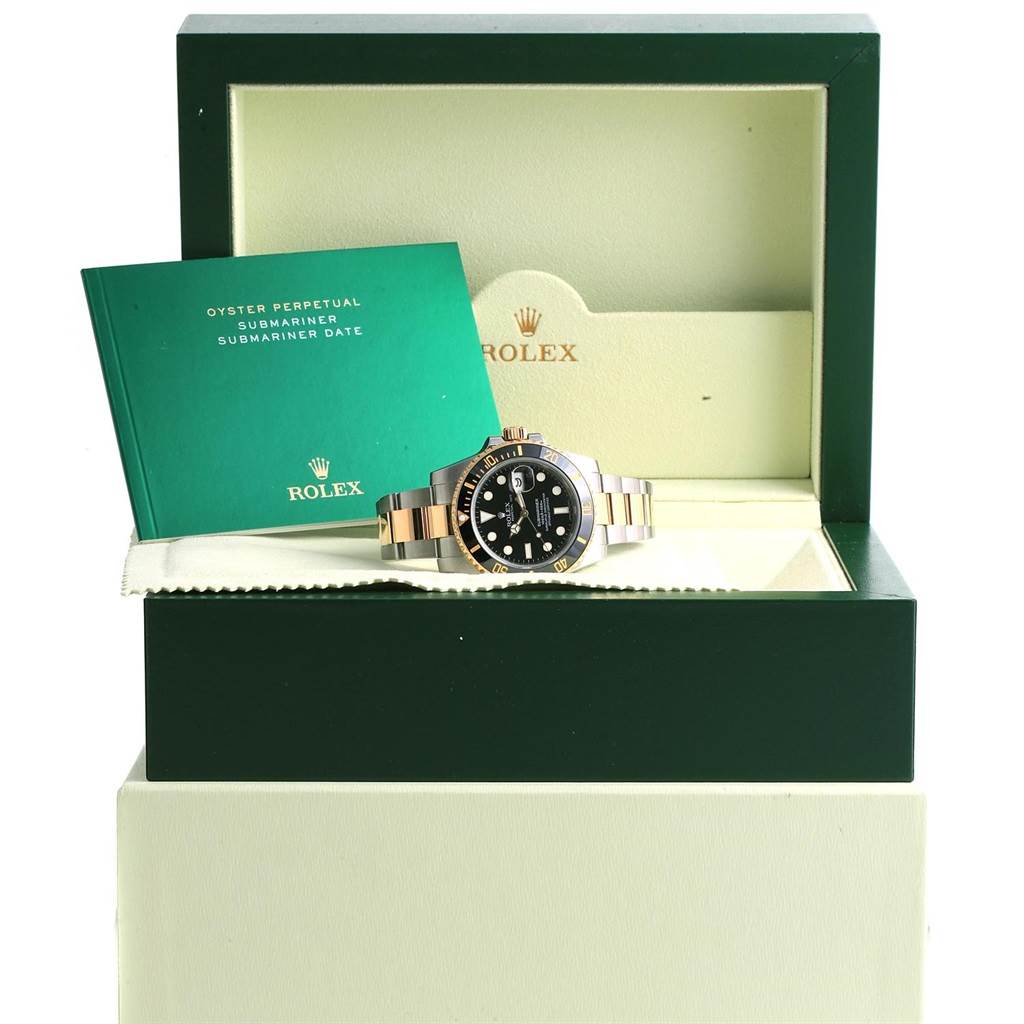 The Rolex Submariner 116613 Men's Steel and Gold (two tone) Black Dial 116613 Men's Steel and Gold (two tone) Black Dial watch is shown in its open box with a green manual beside it, viewed from the front.