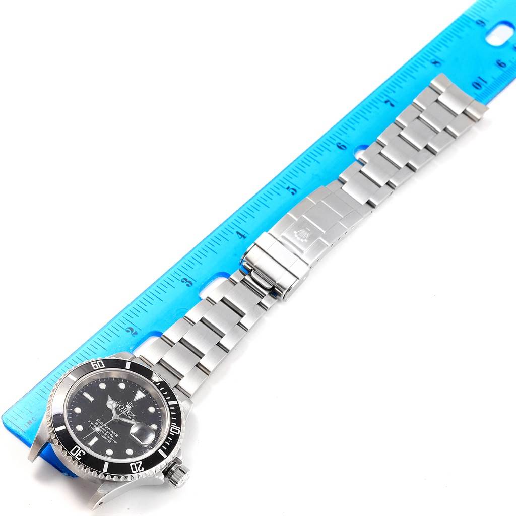 The Rolex Submariner 16610 Men's Stainless Steel Black Dial 16610 Men's Stainless Steel Black Dial watch is shown from a top-down angle, featuring its face, bezel, and steel bracelet on a blue ruler.