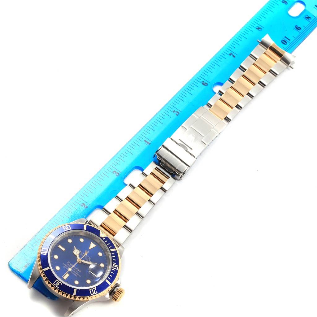 This image shows a Rolex Submariner 16613 Men's Steel and Gold (two tone) Blue Dial 16613 Men's Steel and Gold (two tone) Blue Dial watch positioned next to a ruler, highlighting its blue dial, bezel, and two-tone bracelet.