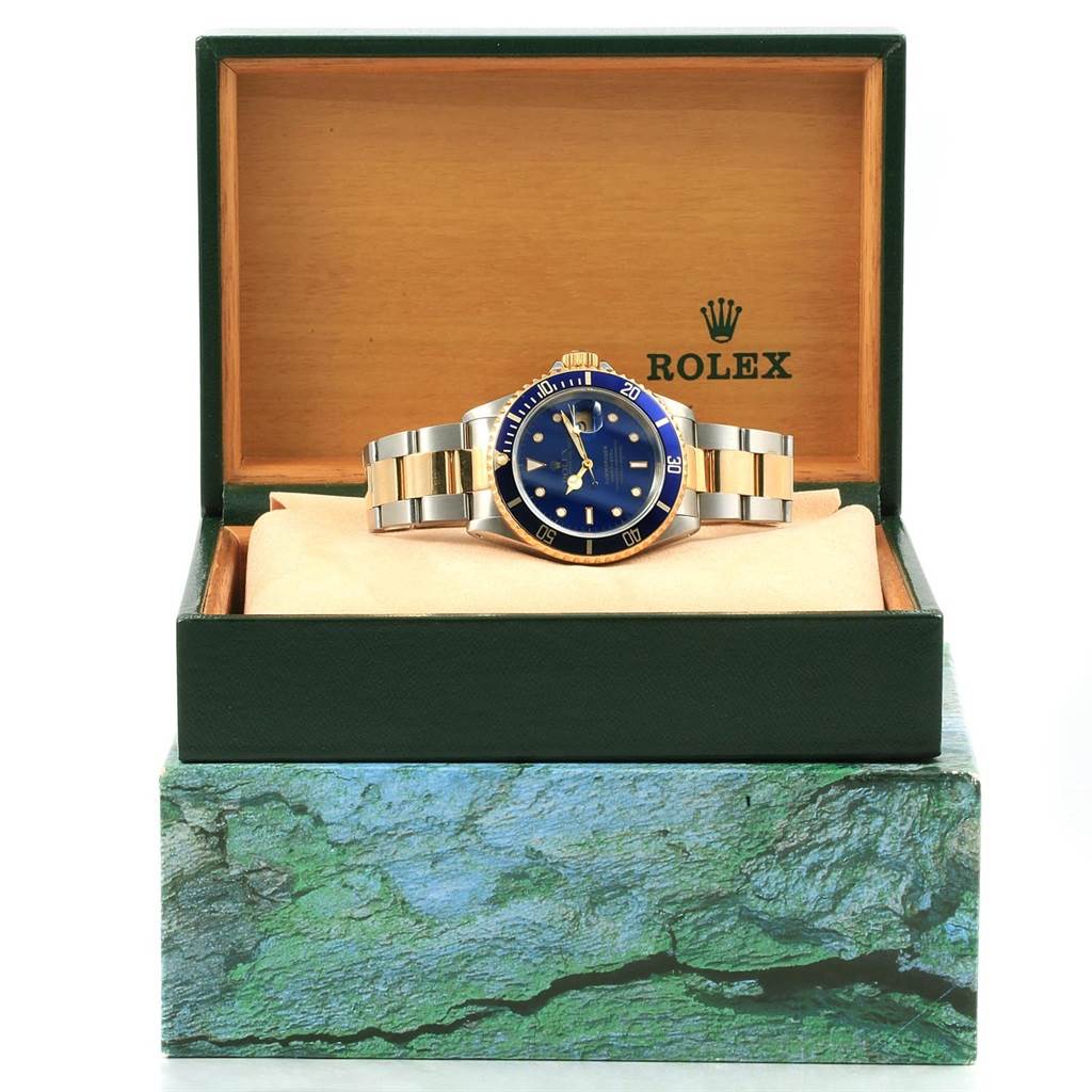 The Rolex Submariner 16613 Men's Steel and Gold (two tone) Blue Dial 16613 Men's Steel and Gold (two tone) Blue Dial watch is shown front-facing in its open green and wood-accented box.