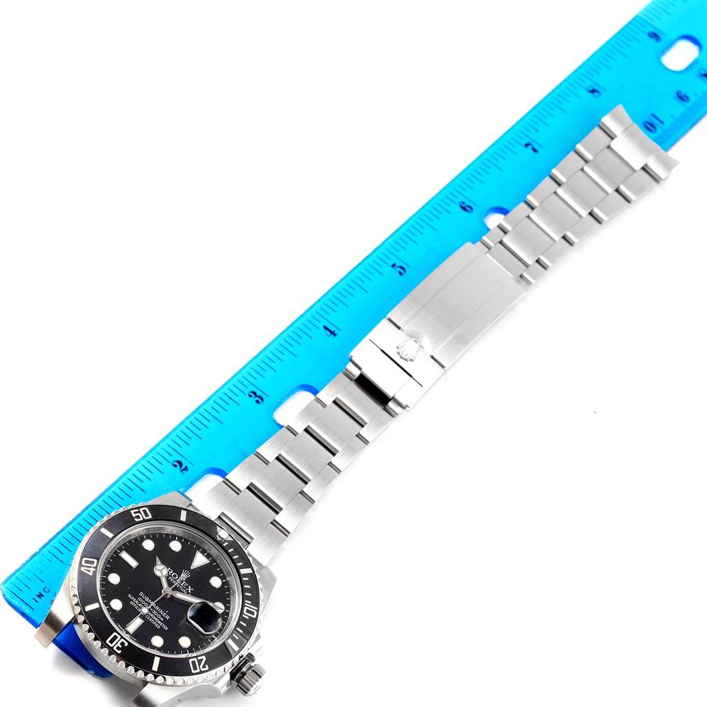 The image shows a Rolex Submariner 116610 Men
s Stainless Steel Black Dial 116610 Men
s Stainless Steel Black Dial watch laid flat with a blue ruler beside it, displaying the watch face and stainless steel bracelet.