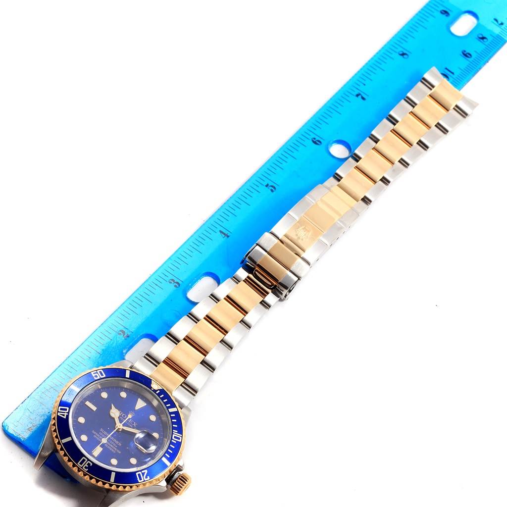 The image shows a Rolex Submariner 16613 Men
s Steel and Gold (two tone) Blue Dial 16613 Men
s Steel and Gold (two tone) Blue Dial watch placed alongside a ruler, displaying its blue dial, bezel, and two-tone bracelet.