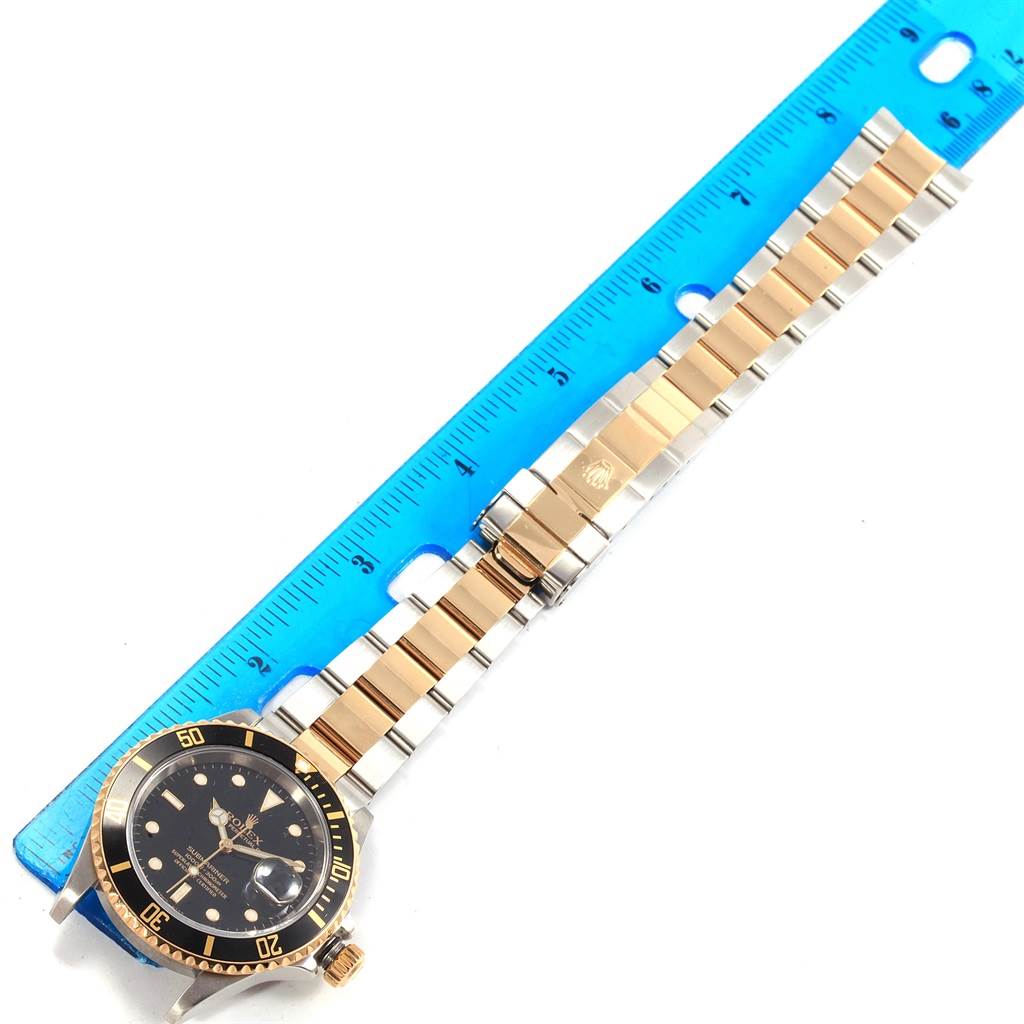 The image shows a Rolex Submariner 16613 Men's Steel and Gold (two tone) Black Dial 16613 Men's Steel and Gold (two tone) Black Dial watch positioned horizontally next to a blue ruler, displaying the face, bezel, and bracelet.