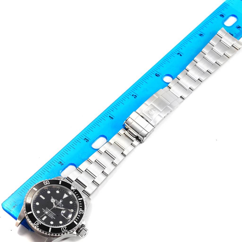 The image shows a Rolex Submariner 16610 Men
s Stainless Steel Black Dial 16610 Men
s Stainless Steel Black Dial watch laid flat next to a ruler, displaying its face and metal bracelet.