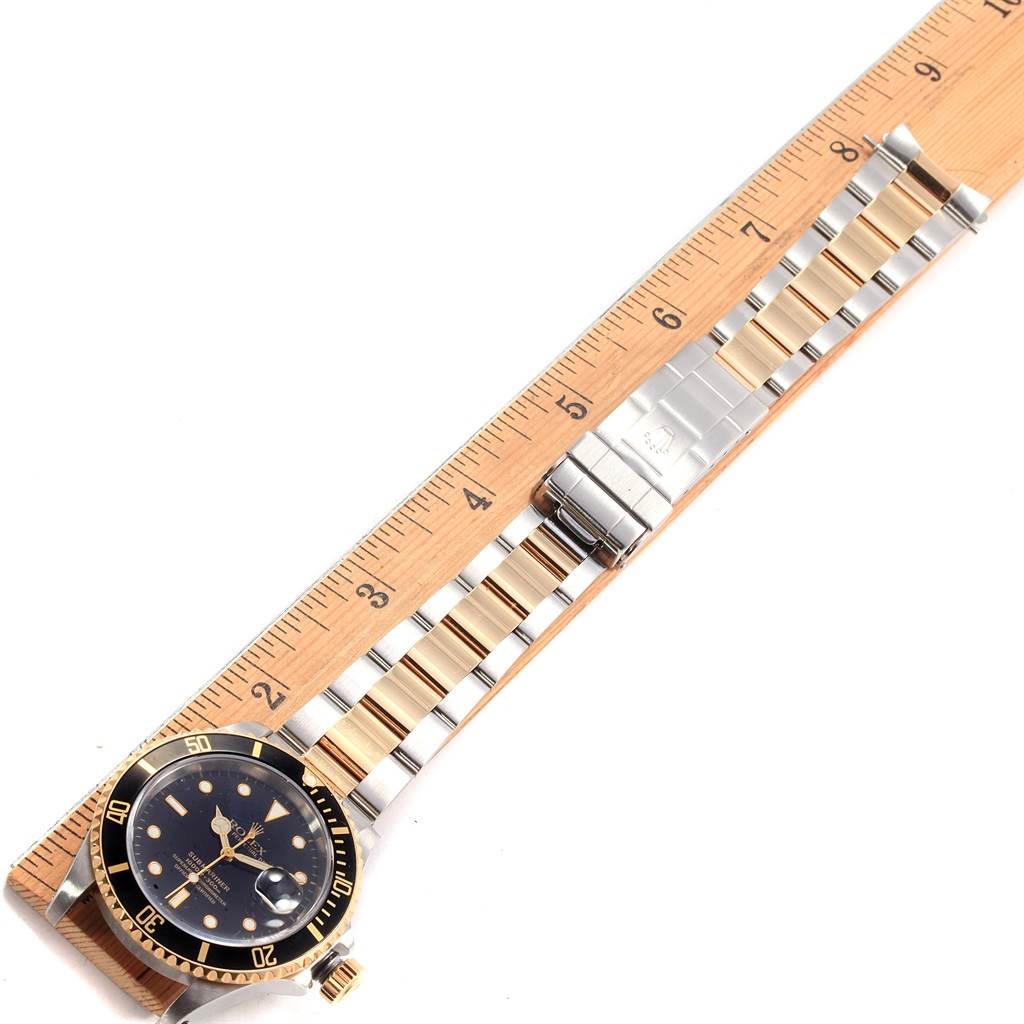 The image shows a Rolex Submariner 16613 Men's Steel and Gold (two tone) Black Dial 16613 Men's Steel and Gold (two tone) Black Dial watch laid flat next to a ruler, highlighting the watch's face and bracelet.