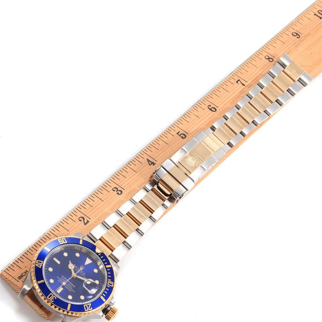 This image shows a Rolex Submariner 16613 Men
s Steel and Gold (two tone) Blue Dial 16613 Men
s Steel and Gold (two tone) Blue Dial watch with a blue dial and bezel, and a two-tone metal bracelet laid flat next to a ruler.