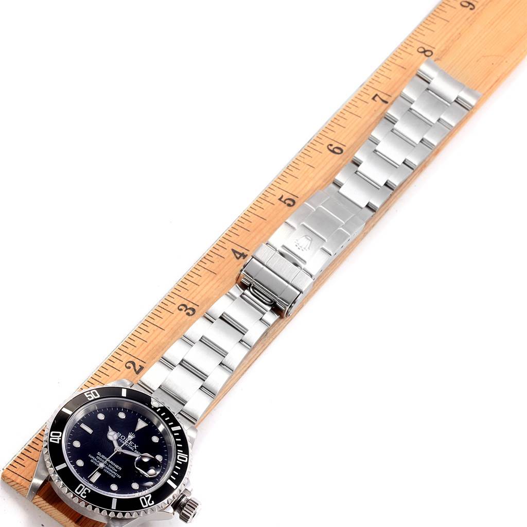 The image shows a Rolex Submariner 16610 Men
s Stainless Steel Black Dial 16610 Men
s Stainless Steel Black Dial watch laid flat against a wooden ruler, displaying its face, bezel, and bracelet with clasp.