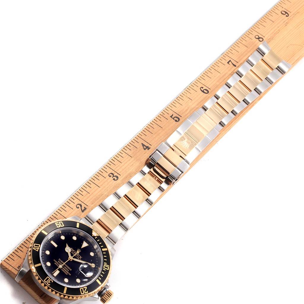 The Rolex Submariner 16613 Men's Steel and Gold (two tone) Black Dial 16613 Men's Steel and Gold (two tone) Black Dial watch is shown from an overhead angle, highlighting the dial, bezel, and metal bracelet alongside a ruler.