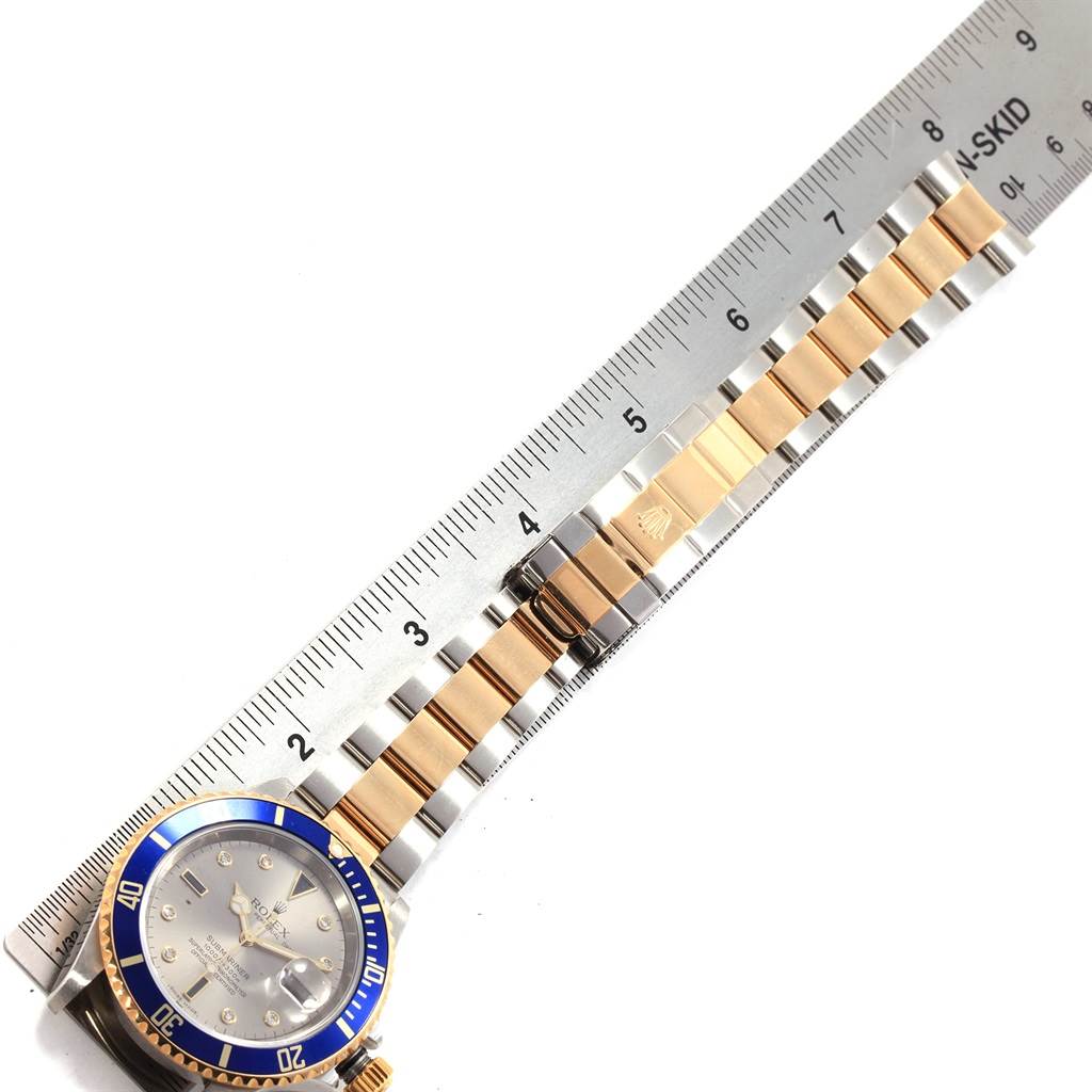 The image shows the Rolex Submariner 16613 Men's Steel and Gold (two tone) Grey Dial 16613 Men's Steel and Gold (two tone) Grey Dial watch at an angle revealing the round dial, bracelet, and part of a ruler for scale.