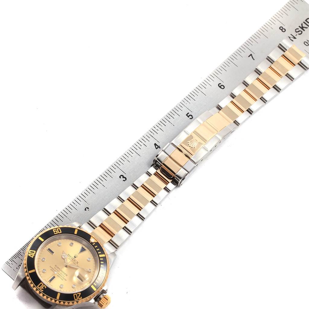The Rolex Submariner 16613 Men's Steel and Gold (two tone) Champagne Dial 16613 Men's Steel and Gold (two tone) Champagne Dial watch is shown laid flat next to a ruler, displaying the dial, bezel, and metal bracelet.