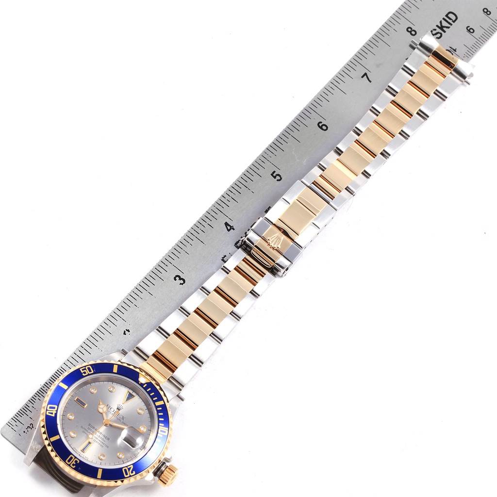 The image shows a Rolex Submariner 16613 Men's Steel and Gold (two tone) Grey Dial 16613 Men's Steel and Gold (two tone) Grey Dial watch with a ruler next to it, highlighting its blue bezel and gold and silver band.