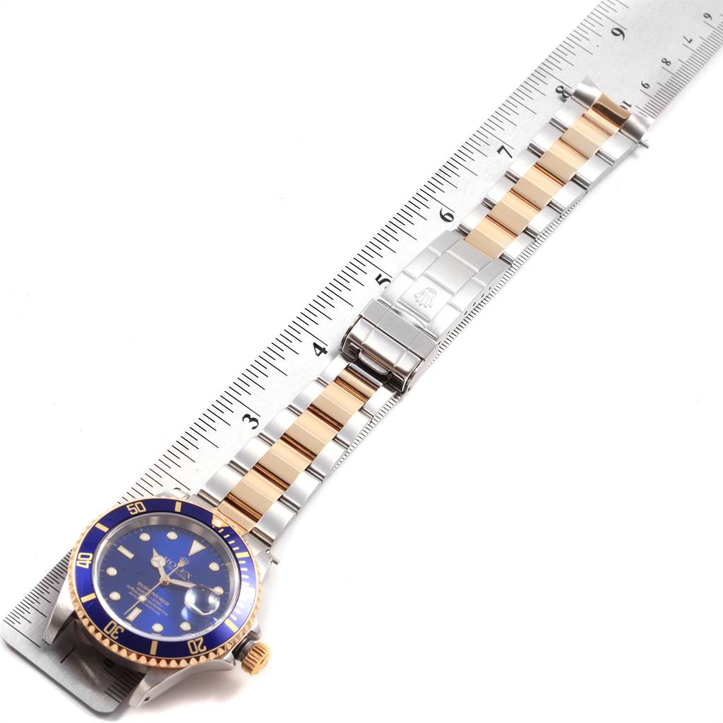 The image shows a Rolex Submariner 16613 Men's Steel and Gold (two tone) Blue Dial 16613 Men's Steel and Gold (two tone) Blue Dial watch at an angle, displaying its face, bezel, and two-tone metal bracelet next to a ruler.