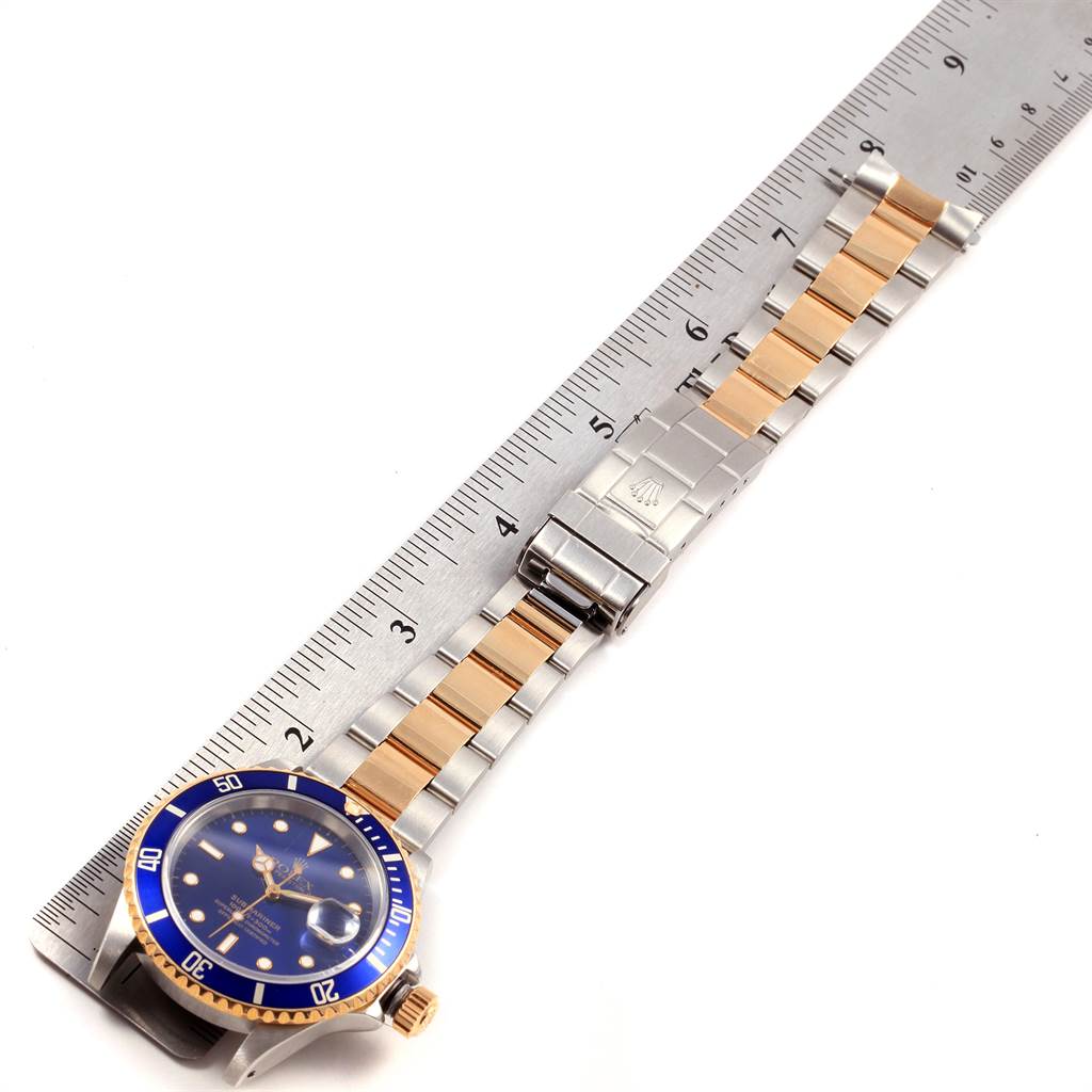 The Rolex Submariner 16613 Men's Steel and Gold (two tone) Blue Dial 16613 Men's Steel and Gold (two tone) Blue Dial watch is shown laid flat next to a ruler, displaying its blue dial, gold accents, and metal bracelet.