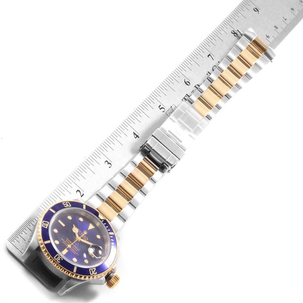 The image shows a Rolex Submariner 16613 Men's Steel and Gold (two tone) Blue Dial 16613 Men's Steel and Gold (two tone) Blue Dial watch with a blue dial, gold bezel, and a two-tone metal band lying alongside a ruler.