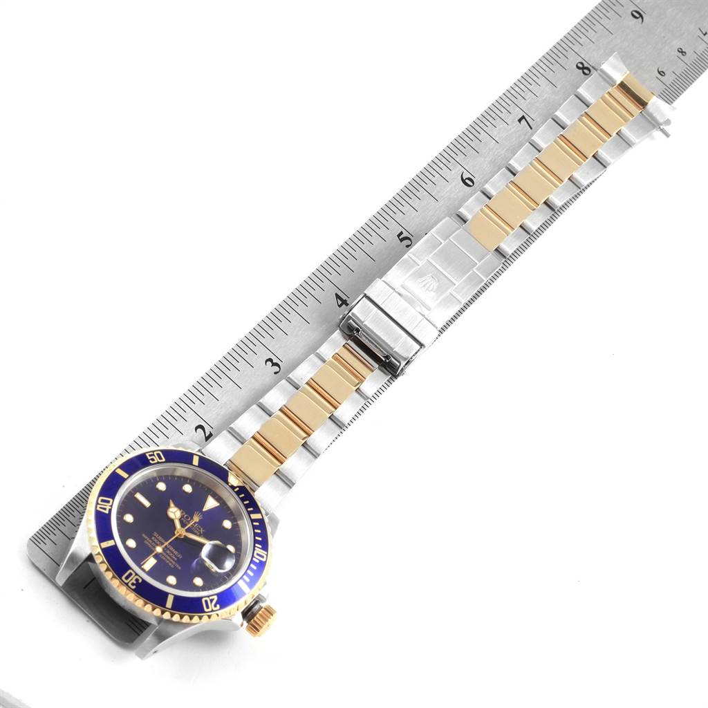 The image shows a top view of a Rolex Submariner 16613 Men's Steel and Gold (two tone) Blue Dial 16613 Men's Steel and Gold (two tone) Blue Dial watch with a blue dial and two-tone metal bracelet, laid next to a ruler.