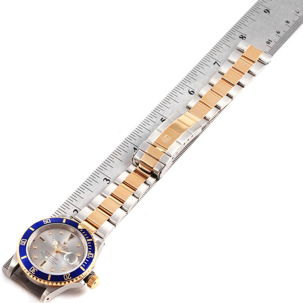 The image shows a Rolex Submariner 16613 Men's Steel and Gold (two tone) Grey Dial 16613 Men's Steel and Gold (two tone) Grey Dial watch laid flat, highlighting its blue bezel, face, two-tone bracelet, and clasp alongside a ruler.