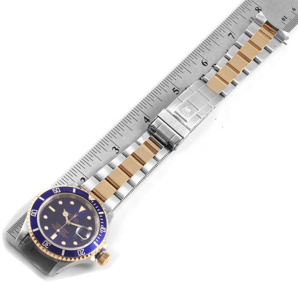 The image shows a Rolex Submariner 16613 Men's Steel and Gold (two tone) Blue Dial 16613 Men's Steel and Gold (two tone) Blue Dial watch laid flat with a ruler, displaying its band, clasp, and face.