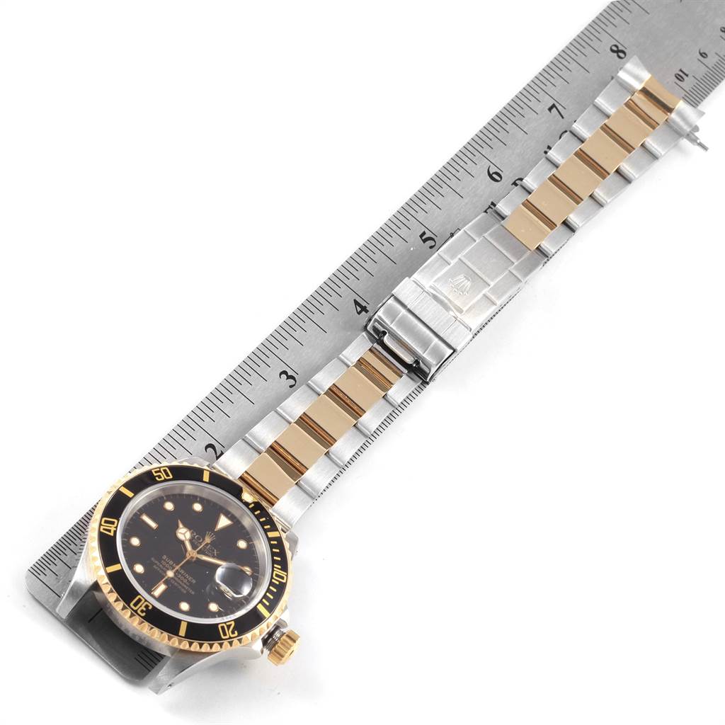 The image shows a Rolex Submariner 16613 Men's Steel and Gold (two tone) Black Dial 16613 Men's Steel and Gold (two tone) Black Dial watch laid flat on a ruler, displaying the dial, bezel, and bracelet.