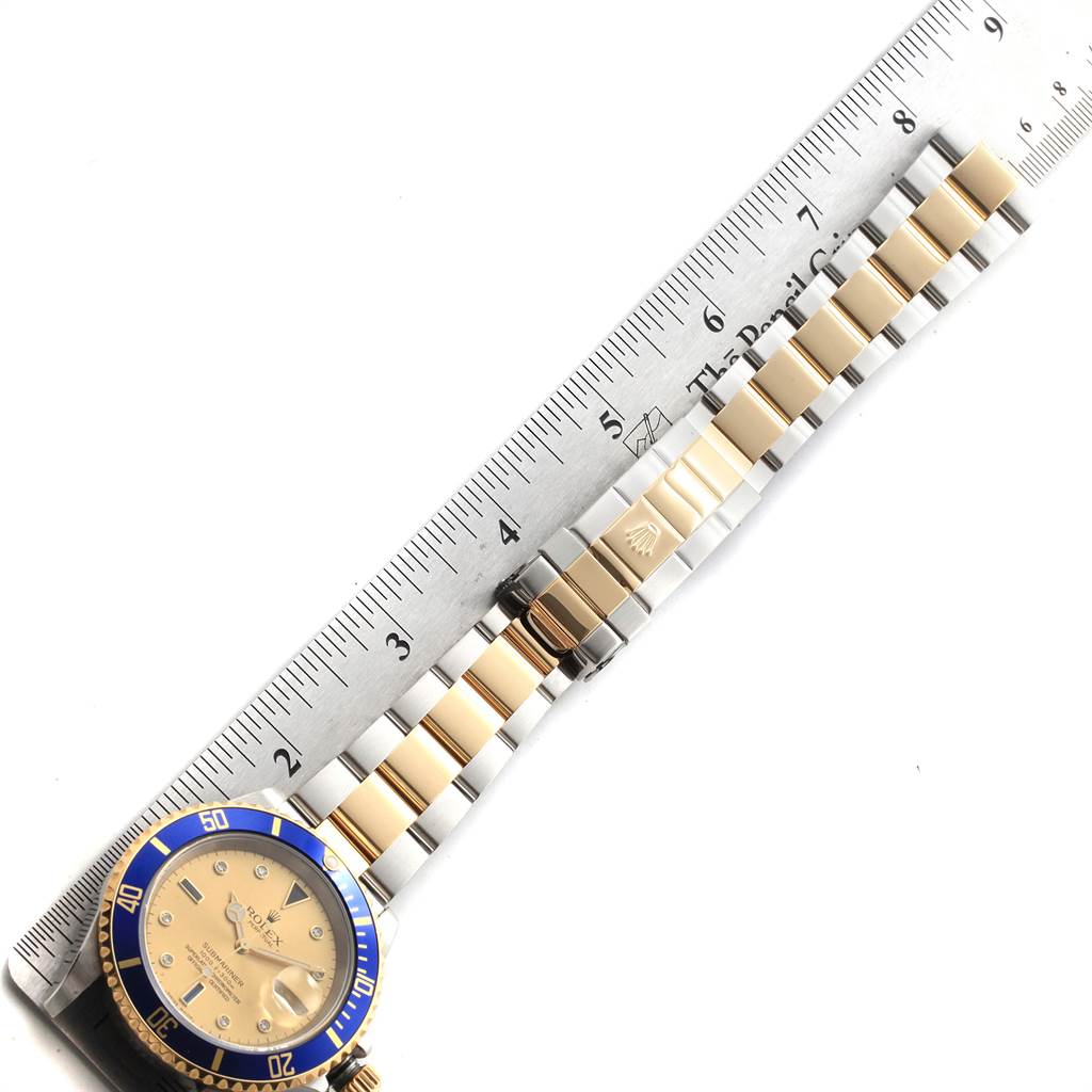 The image shows a top-down view of a Rolex Submariner 16613 Men
s Steel and Gold (two tone) Champagne Dial 16613 Men
s Steel and Gold (two tone) Champagne Dial watch, displaying its blue bezel, gold and silver bracelet, and clasp next to a ruler.