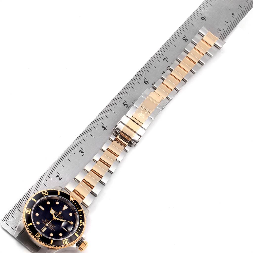 The image shows a Rolex Submariner 16613 Men
s Steel and Gold (two tone) Black Dial 16613 Men
s Steel and Gold (two tone) Black Dial watch with its band extended next to a ruler, displaying the dial and bracelet.