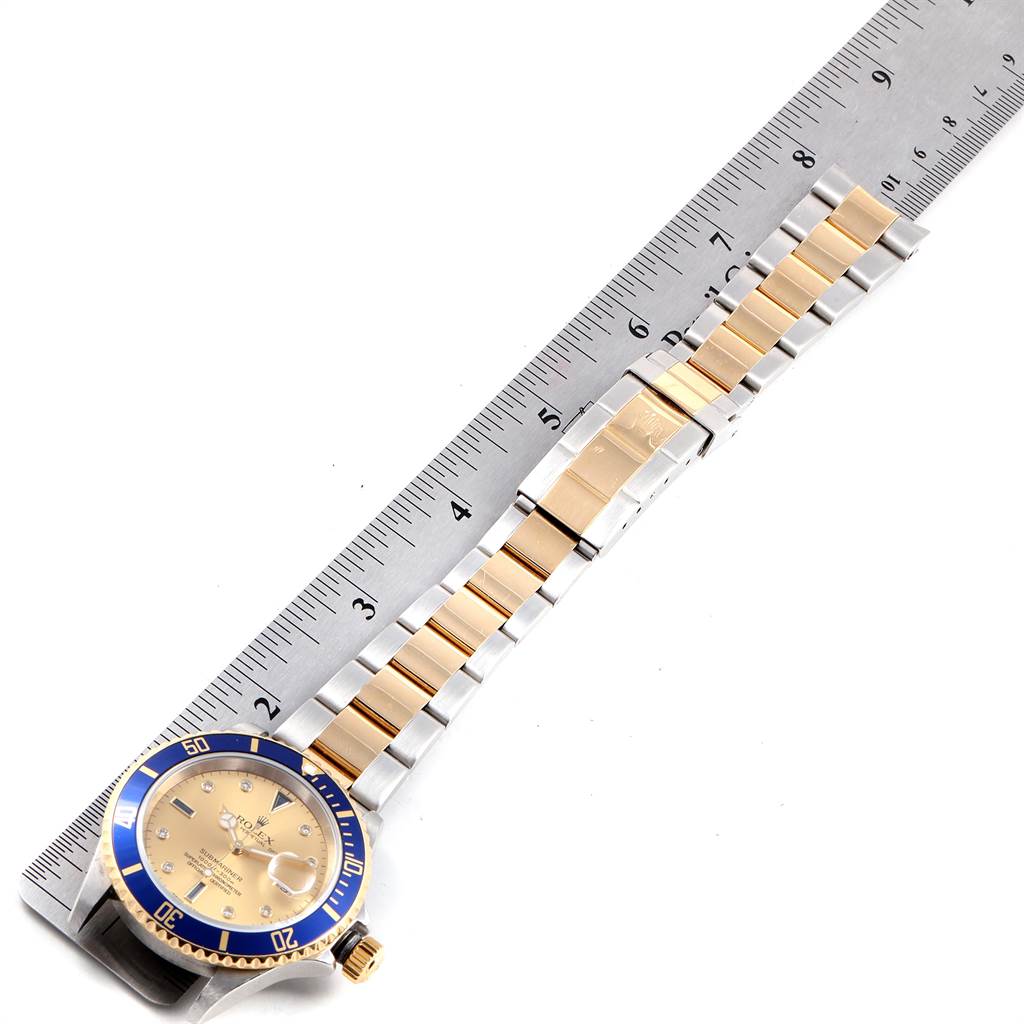 The image shows a top-down view of a Rolex Submariner 16613 Men's Steel and Gold (two tone) Champagne Dial 16613 Men's Steel and Gold (two tone) Champagne Dial watch with a blue bezel and gold-and-silver bracelet, next to a ruler.