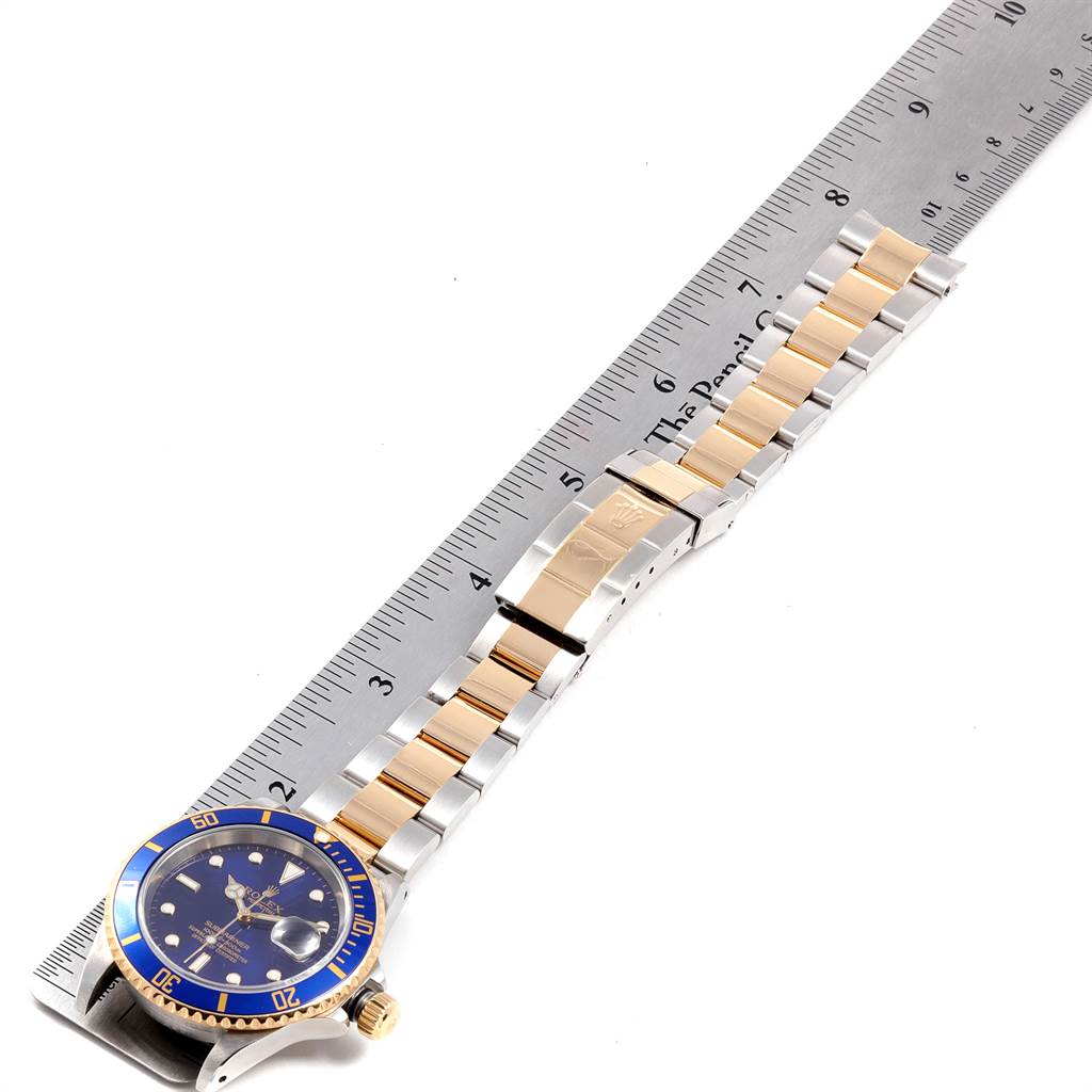The image shows a Rolex Submariner 16613 Men
s Steel and Gold (two tone) Blue Dial 16613 Men
s Steel and Gold (two tone) Blue Dial watch laid flat against a ruler, displaying the watch face, bezel, crown, and band.