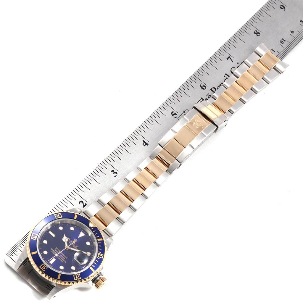 The image shows a Rolex Submariner 16613 Men's Steel and Gold (two tone) Blue Dial 16613 Men's Steel and Gold (two tone) Blue Dial watch, angled diagonally, displaying the dial, bezel, and full bracelet alongside a ruler.