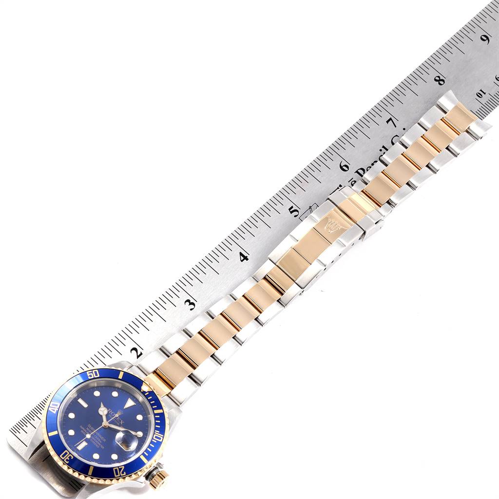 The image shows a top view of a Rolex Submariner 16613 Men's Steel and Gold (two tone) Blue Dial 16613 Men's Steel and Gold (two tone) Blue Dial watch with a blue dial and gold accents alongside a ruler.