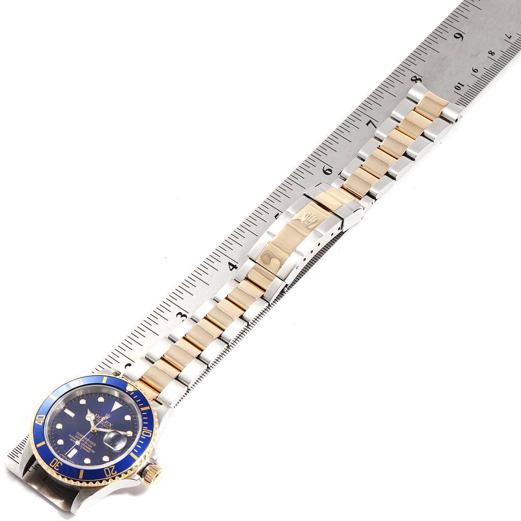 The Rolex Submariner 16613 Men
s Steel and Gold (two tone) Blue Dial 16613 Men
s Steel and Gold (two tone) Blue Dial watch is shown face-up alongside a ruler, displaying the dial, bezel, bracelet, and clasp.