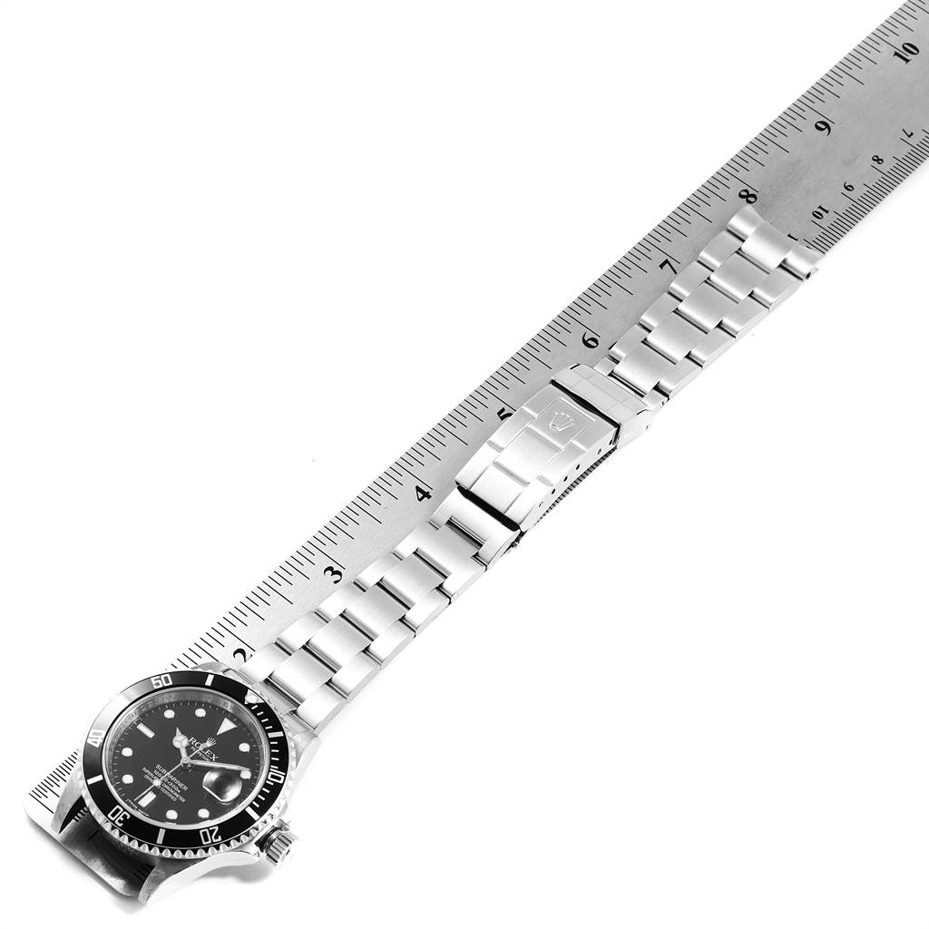 The image shows a Rolex Submariner 16610 Men
s Stainless Steel Black Dial 16610 Men
s Stainless Steel Black Dial watch laid flat next to a metal ruler, displaying the watch face and stainless steel bracelet.