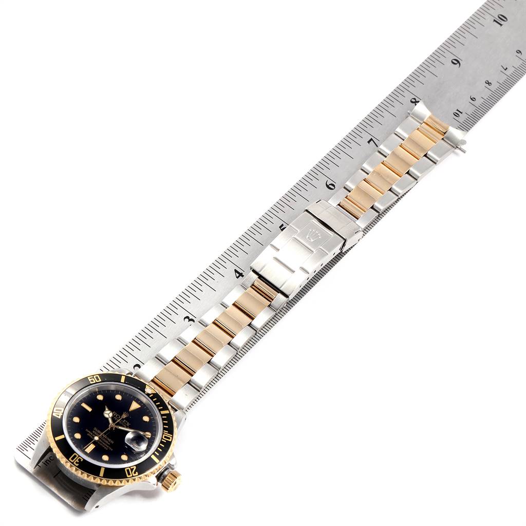The image shows a Rolex Submariner 16803 Men
s Steel and Gold (two tone) Black Dial 16803 Men
s Steel and Gold (two tone) Black Dial watch laid flat against a ruler, highlighting the watch face, bezel, and two-tone bracelet.