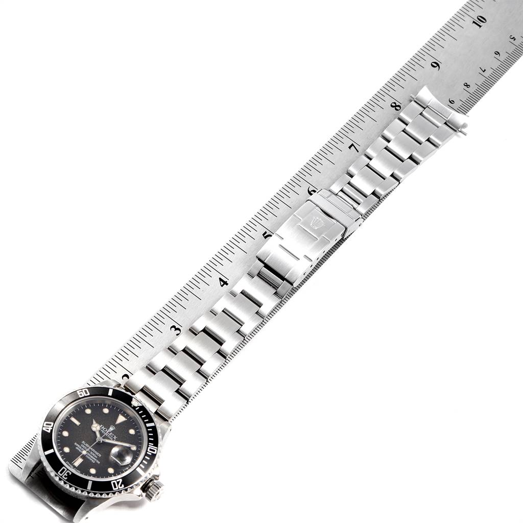 This image shows a Rolex Submariner 16800 Men
s Stainless Steel Black Dial 16800 Men
s Stainless Steel Black Dial watch laid flat, beside a ruler, displaying its face, bezel, bracelet, and clasp.