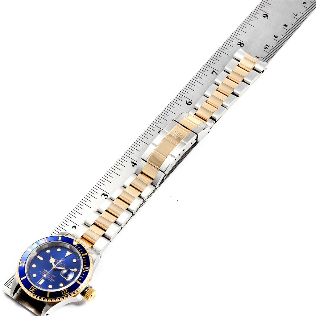 This image shows a Rolex Submariner 16613 Men
s Steel and Gold (two tone) Blue Dial 16613 Men
s Steel and Gold (two tone) Blue Dial watch at an angled top-down view, showcasing its blue dial, bezel, and two-tone bracelet against a ruler.
